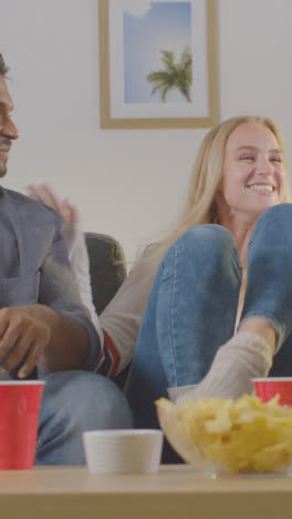 Vertikales-Video-Von-Freunden,-Die-Feiern,-Wie-Sie-Sich-Sportspiele-Im-Fernsehen-Ansehen,-Die-Zu-Hause-Auf-Dem-Sofa-Sitzen-Und-Chips-Und-Dips-Essen